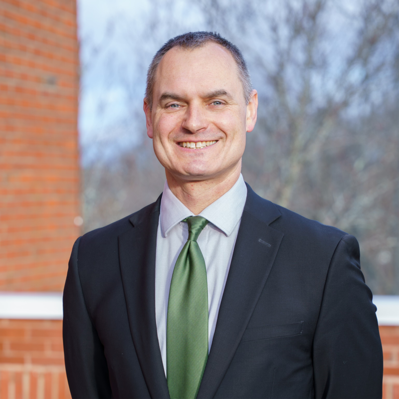 Smiling man in a black suit jacket, light blue shirt, and green tie standing outdoors in front of a brick wall and blurred trees.
