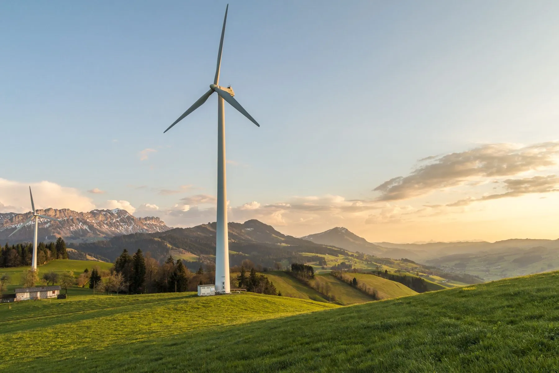 A wind turbine on a green alpine hillside at sunset, snow-capped mountains behind