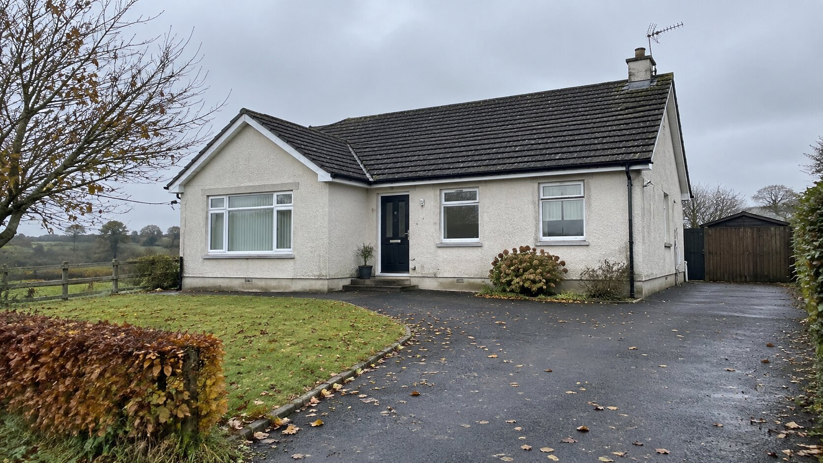 Cream roughcast bungalow with tiled roof, gutters and autumn leaves on a damp driveway.