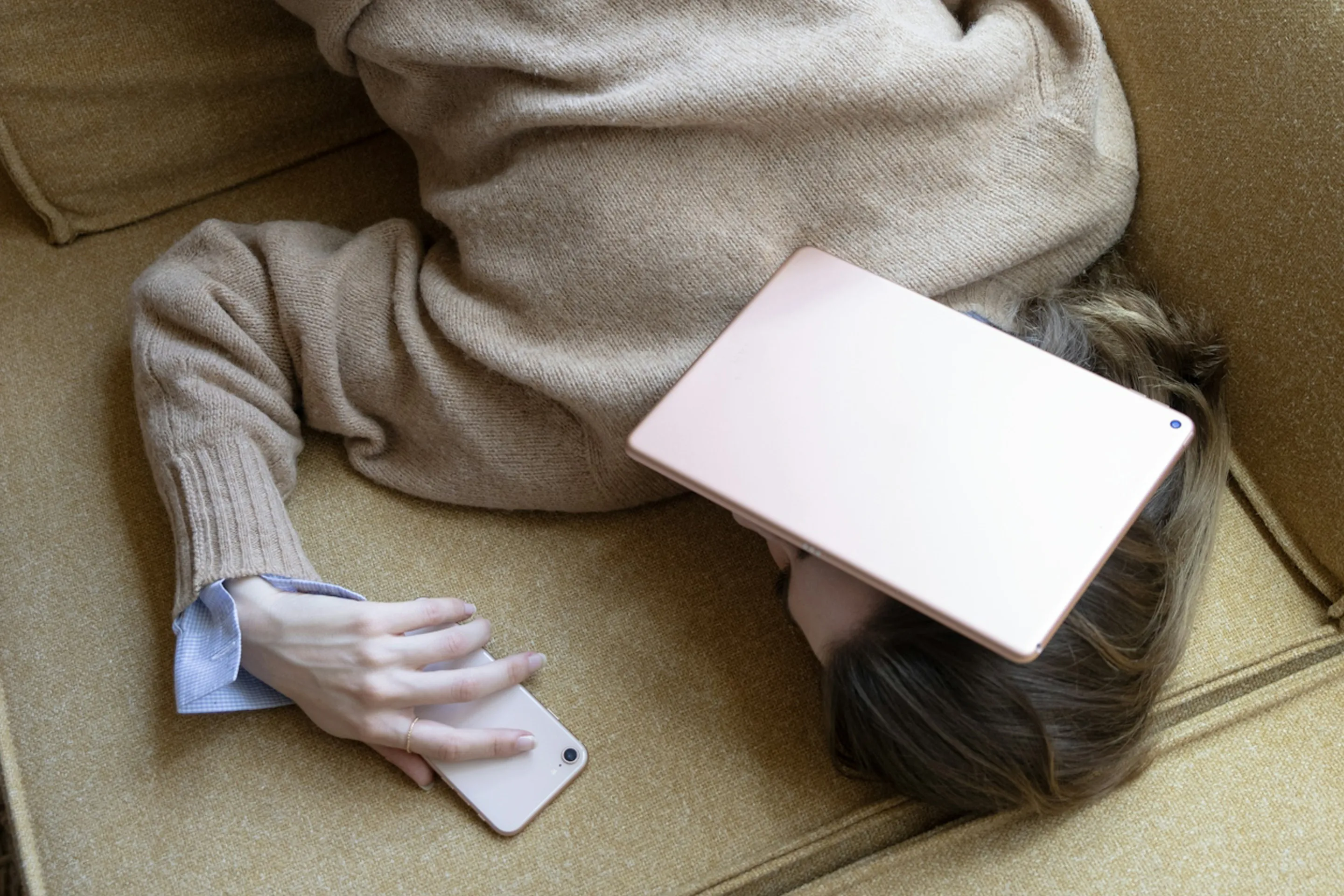 A person lies on a yellow couch, using a smartphone while an electronic tablet rests on their head. They wear a tan sweater and have brown hair, partially visible.
