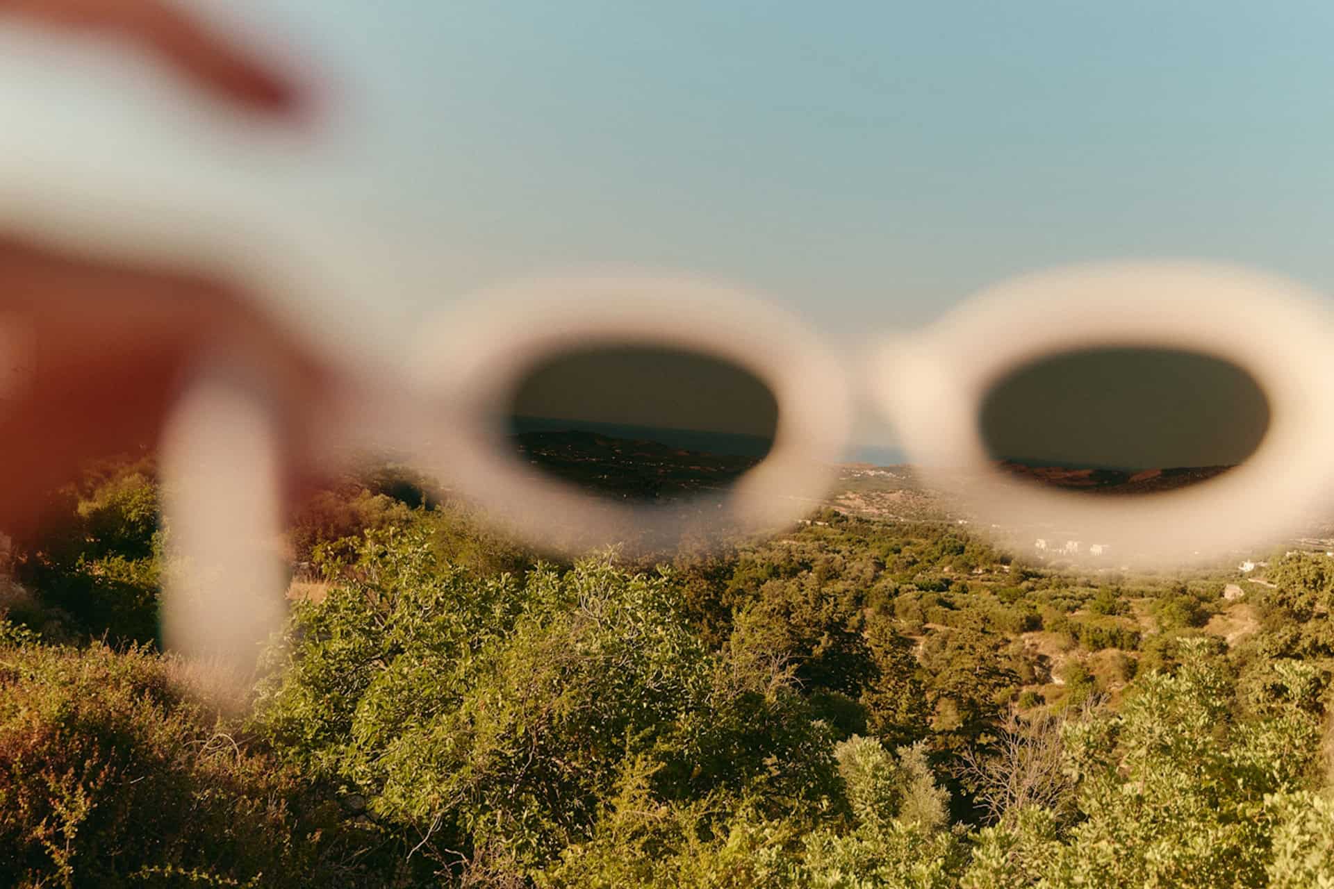 A blurry view of green trees and distant hills, seen through a pair of out-of-focus, round white sunglasses held up in front of a clear blue sky.