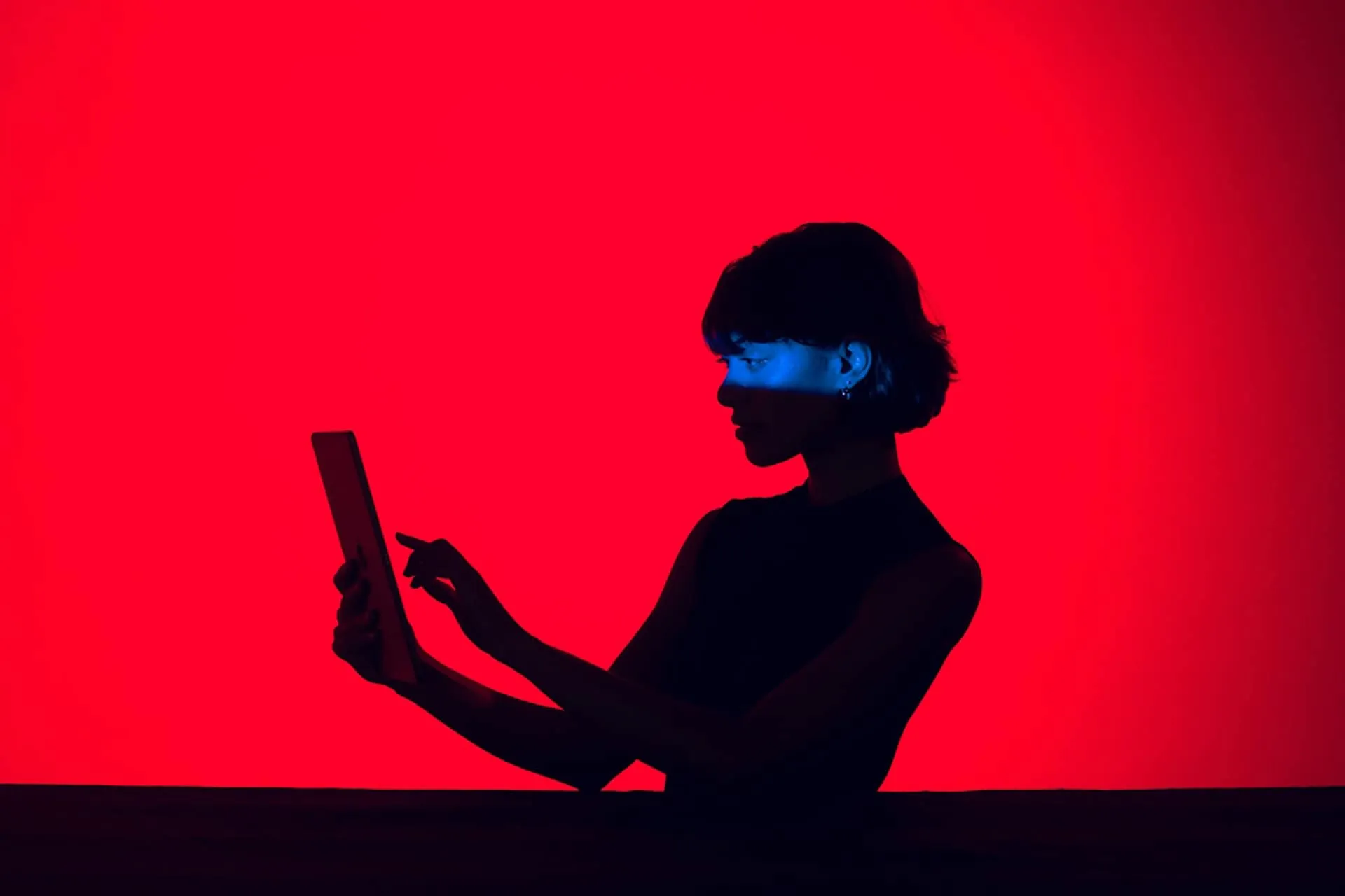 A person in silhouette is interacting with a tablet against a vibrant red background, with blue light illuminating parts of their face and hands.