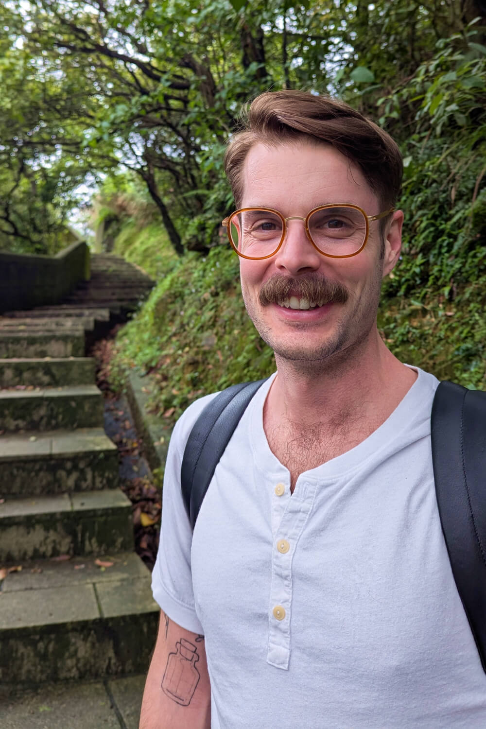 Photo of Alexander in a white shirt standing in front of stone stairs and a lush, leafy background.