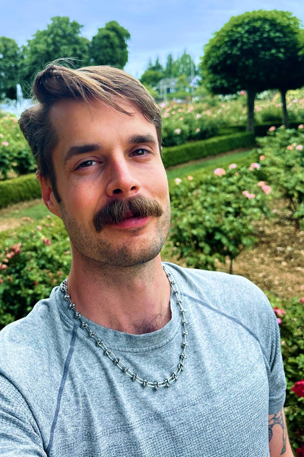 Photo of Alexander in a gray shirt with a metal necklace with a rose garden in the background.
