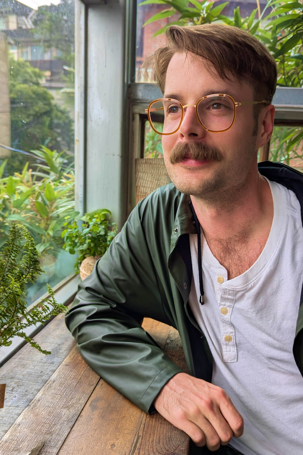 Photo of Alexander wearing brown sunglasses, a white shirt, and a dark green rain jacket at a wooden counter surrounded by glass windows with lush green plants outside.