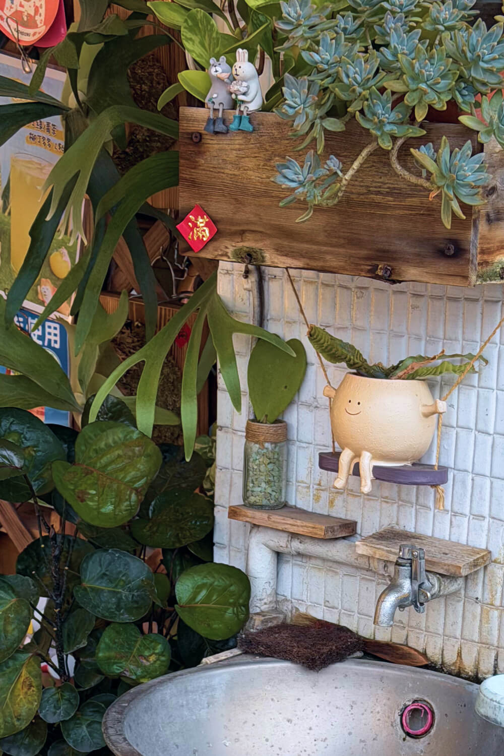A close up scene of a plant in a friendly character pot sitting on a swing above a sink. There are other plants in the background.