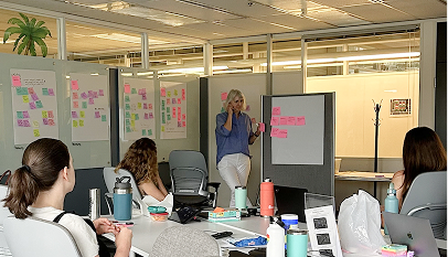A facilitator stands holding sticky notes while presenting to a seated group in an open office space, with whiteboards covered in colorful sticky-note clusters visible in the background.