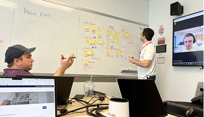 Two team members collaborate around a whiteboard covered in yellow sticky notes mapping a workflow, while a third participant joins via video call on a screen in the background.