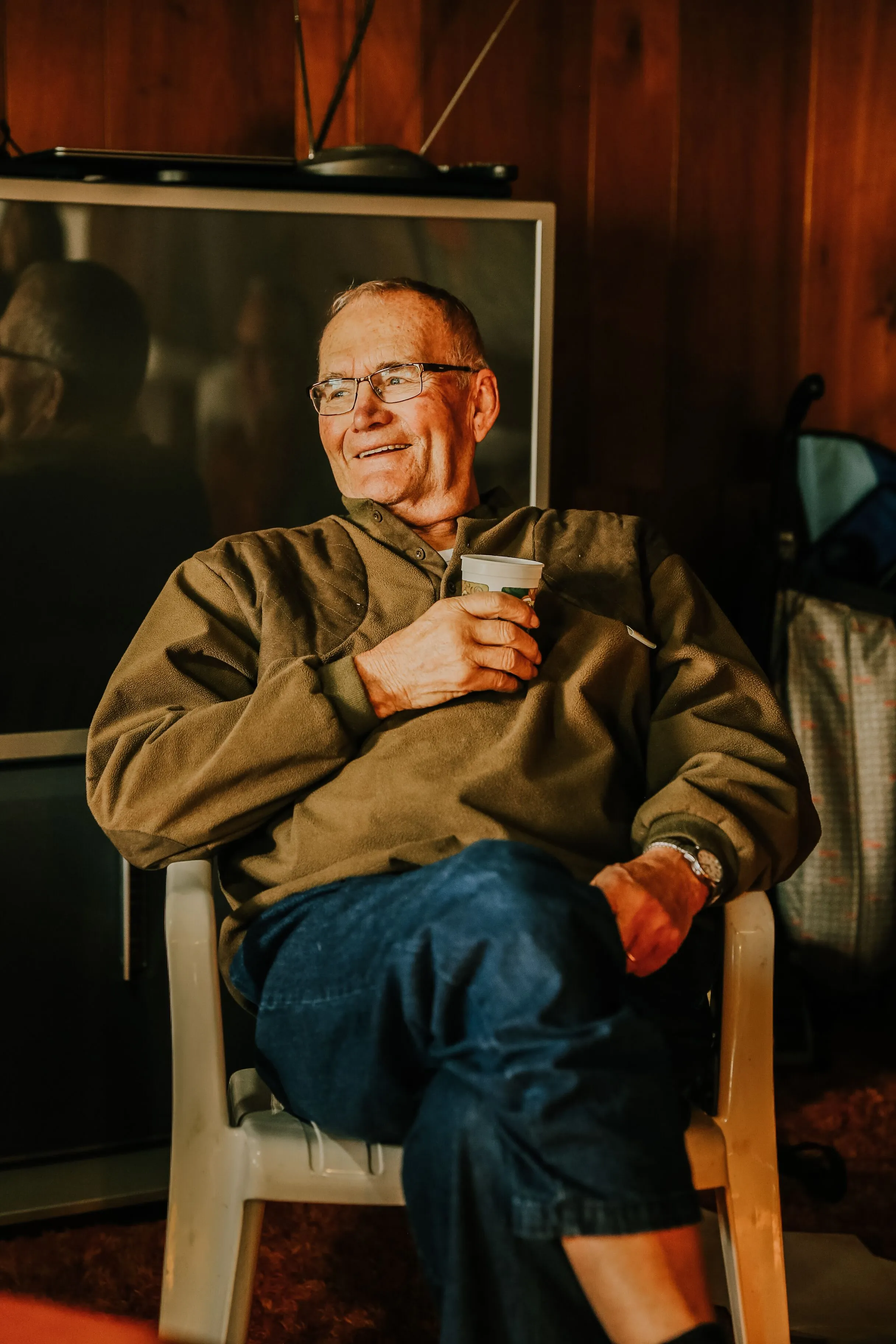 An older man in a fleece pullover relaxing in a chair with a cup of coffee, smiling at someone out of frame