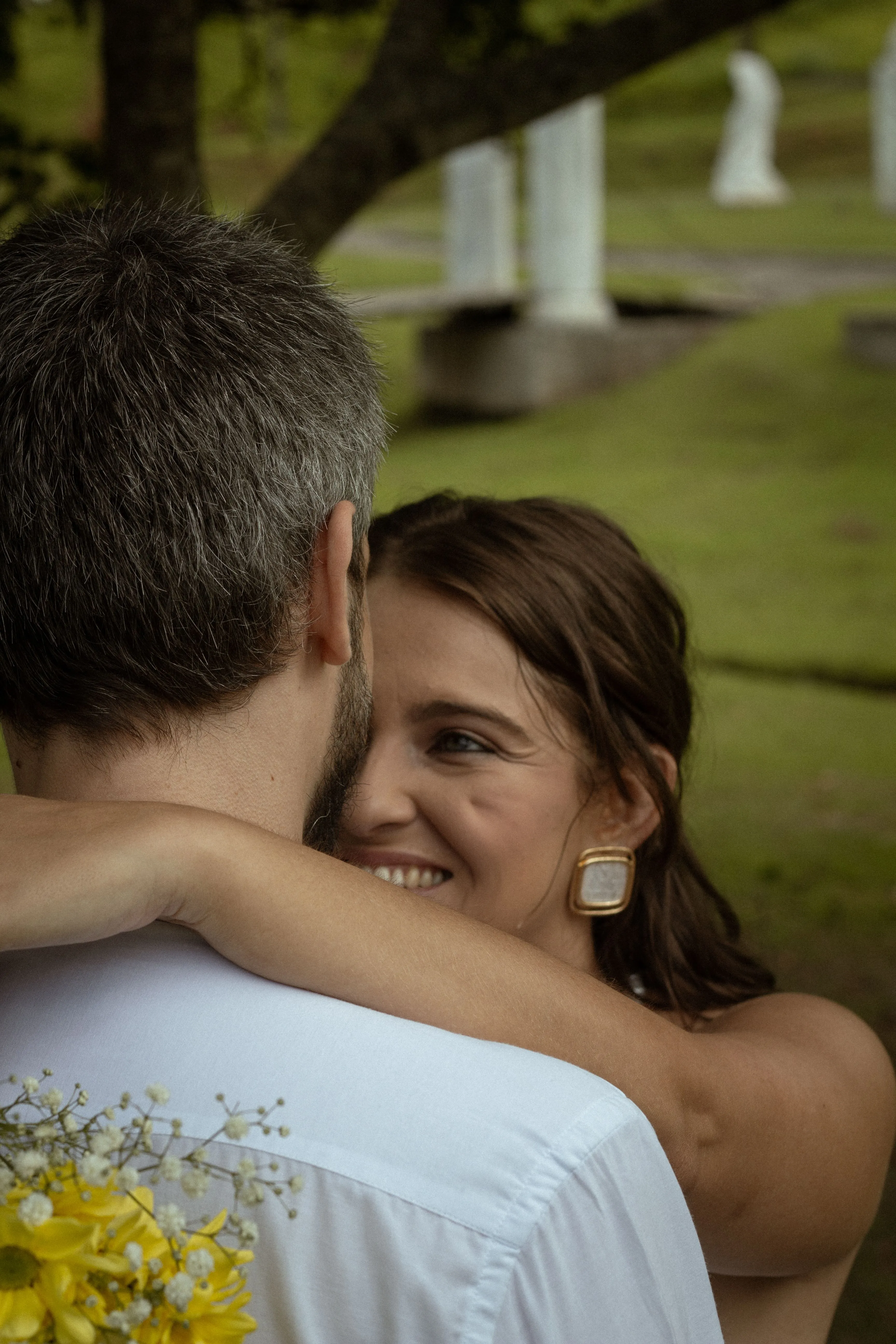 A couple embracing outdoors, her face resting on his shoulder with a quiet smile