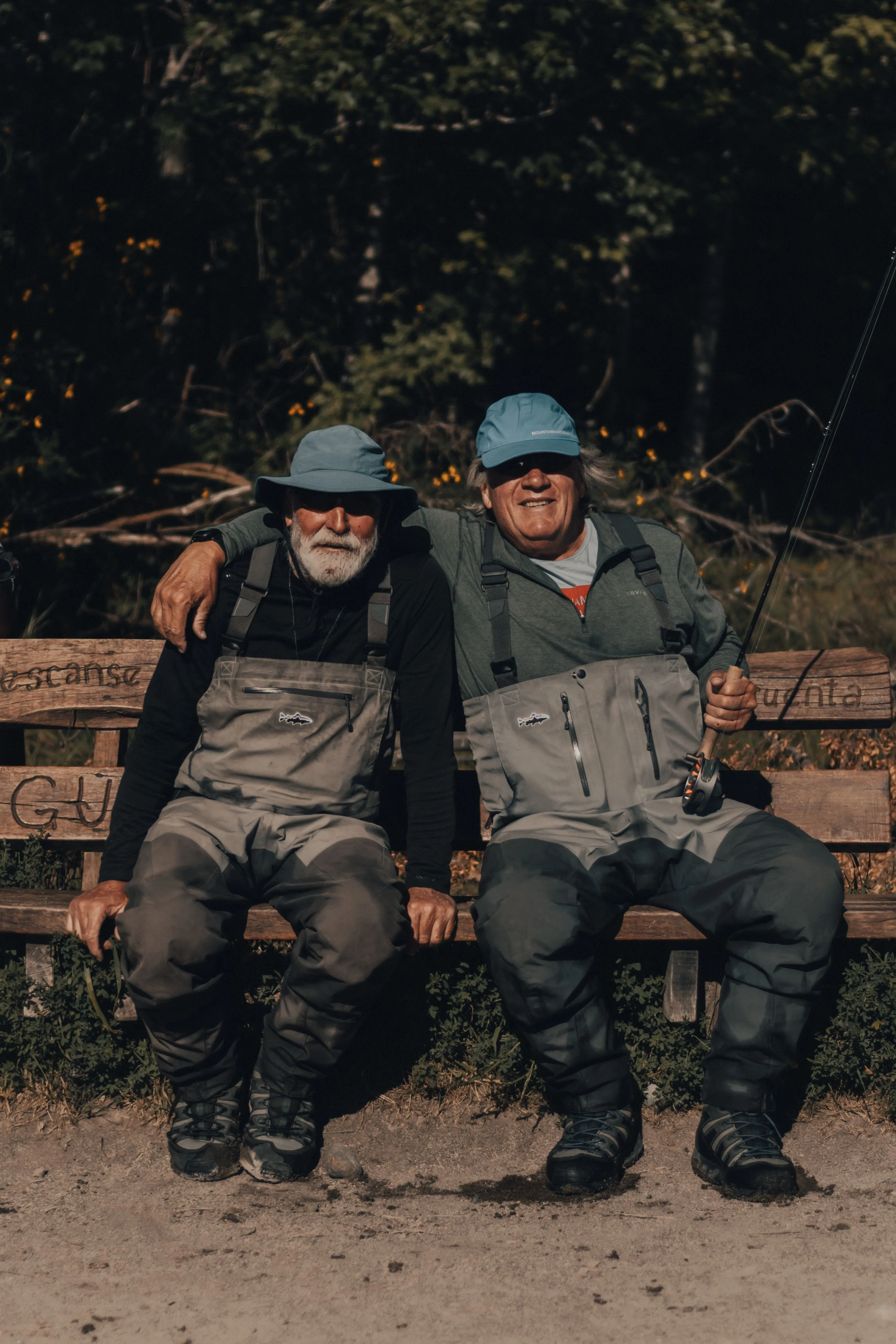 Two older friends in fishing waders sitting on a bench, one with an arm around the other