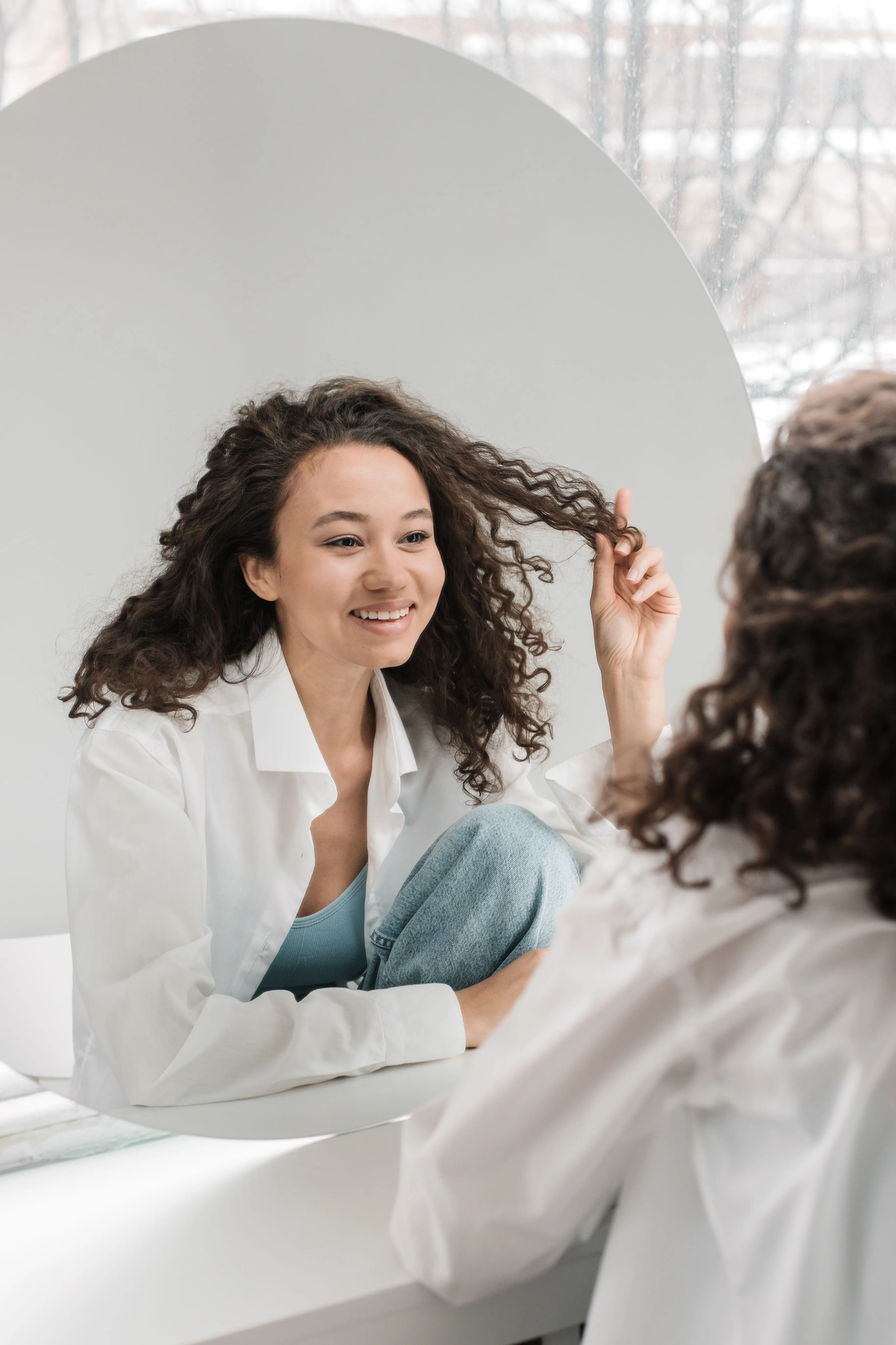 A woman smiling at herself in a round mirror while fixing her hair