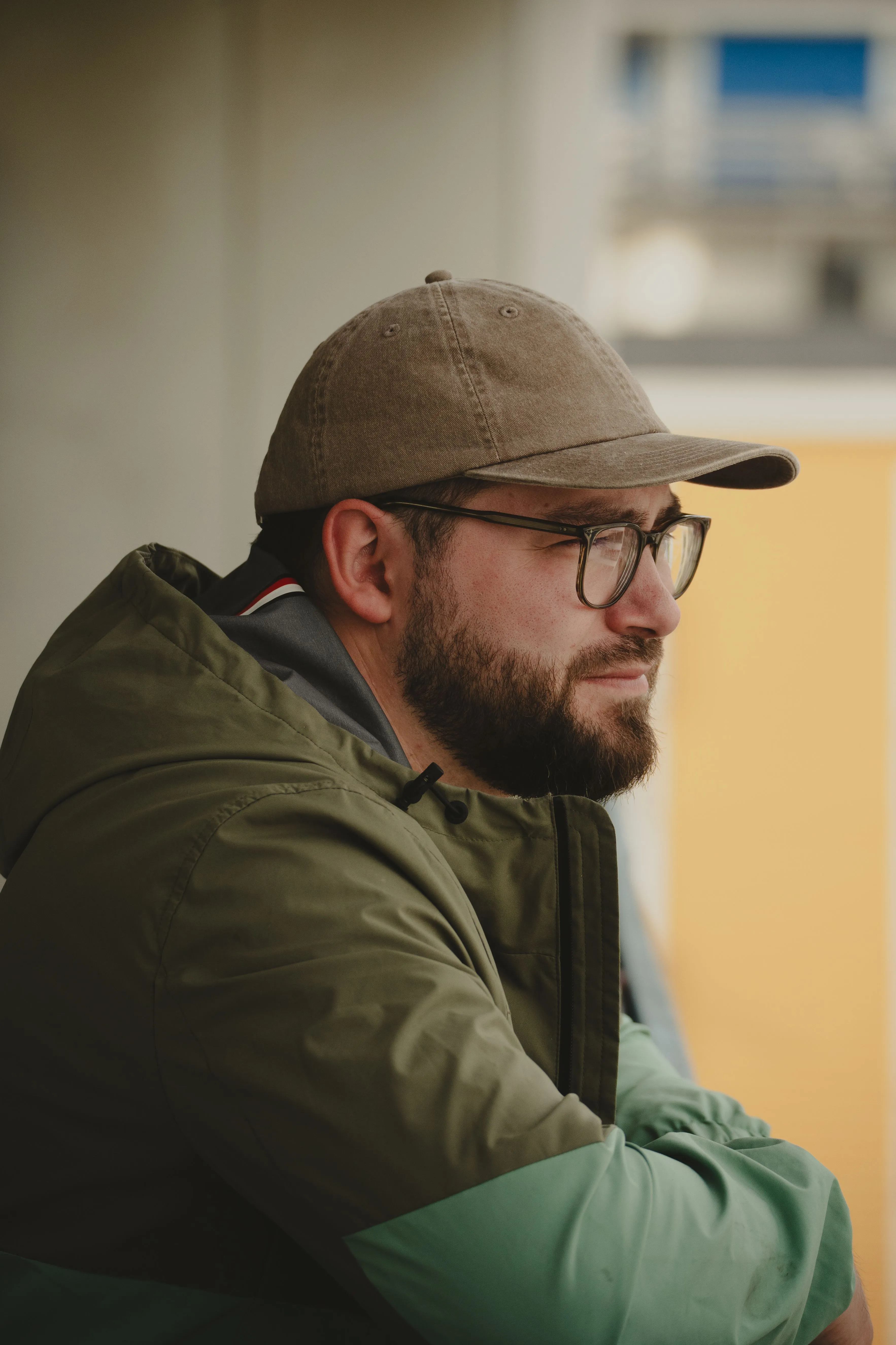 A man in a cap and glasses sitting outside with his arms folded, looking off into the distance