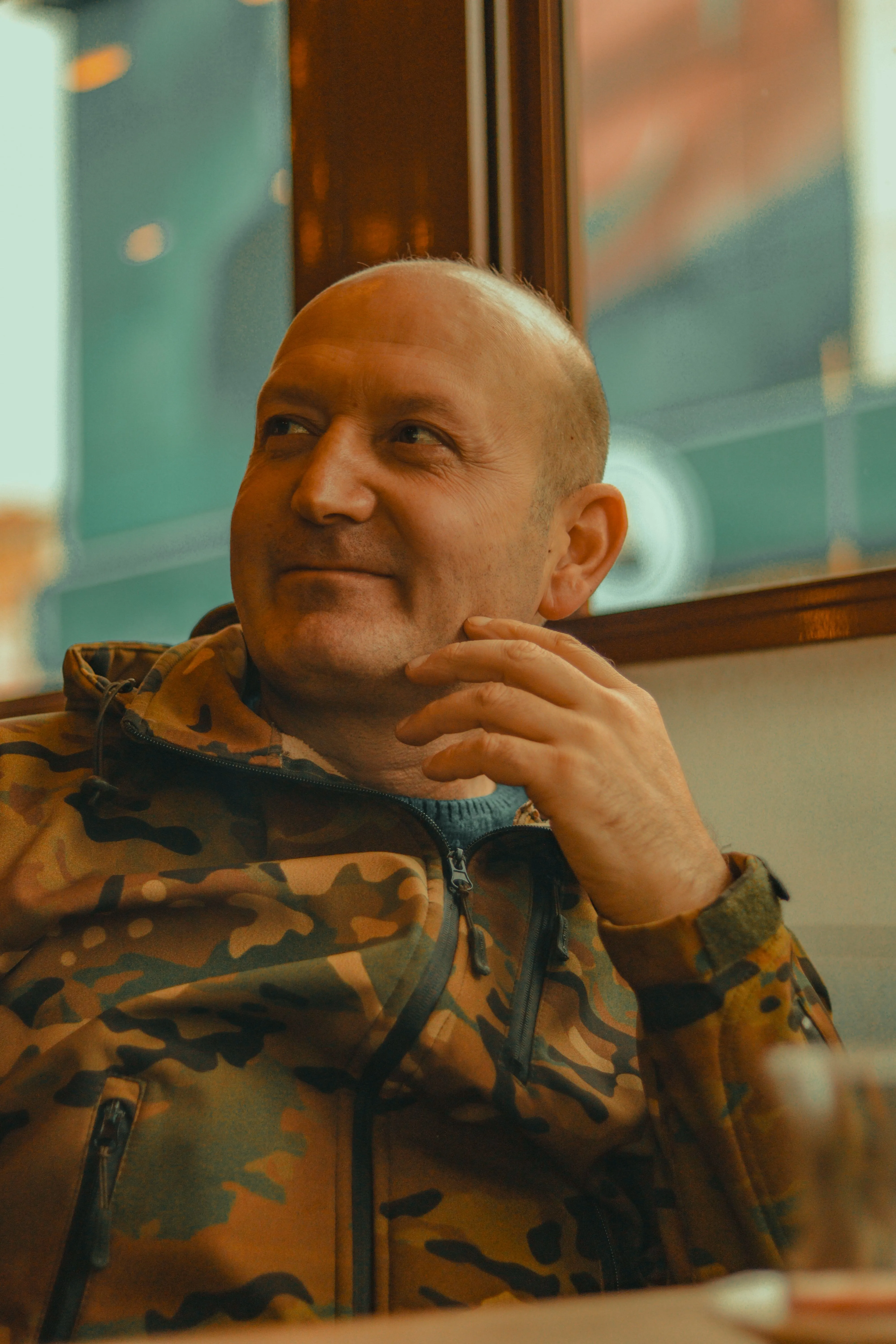 A man in a camouflage jacket sitting in a diner booth, smiling at something across the table