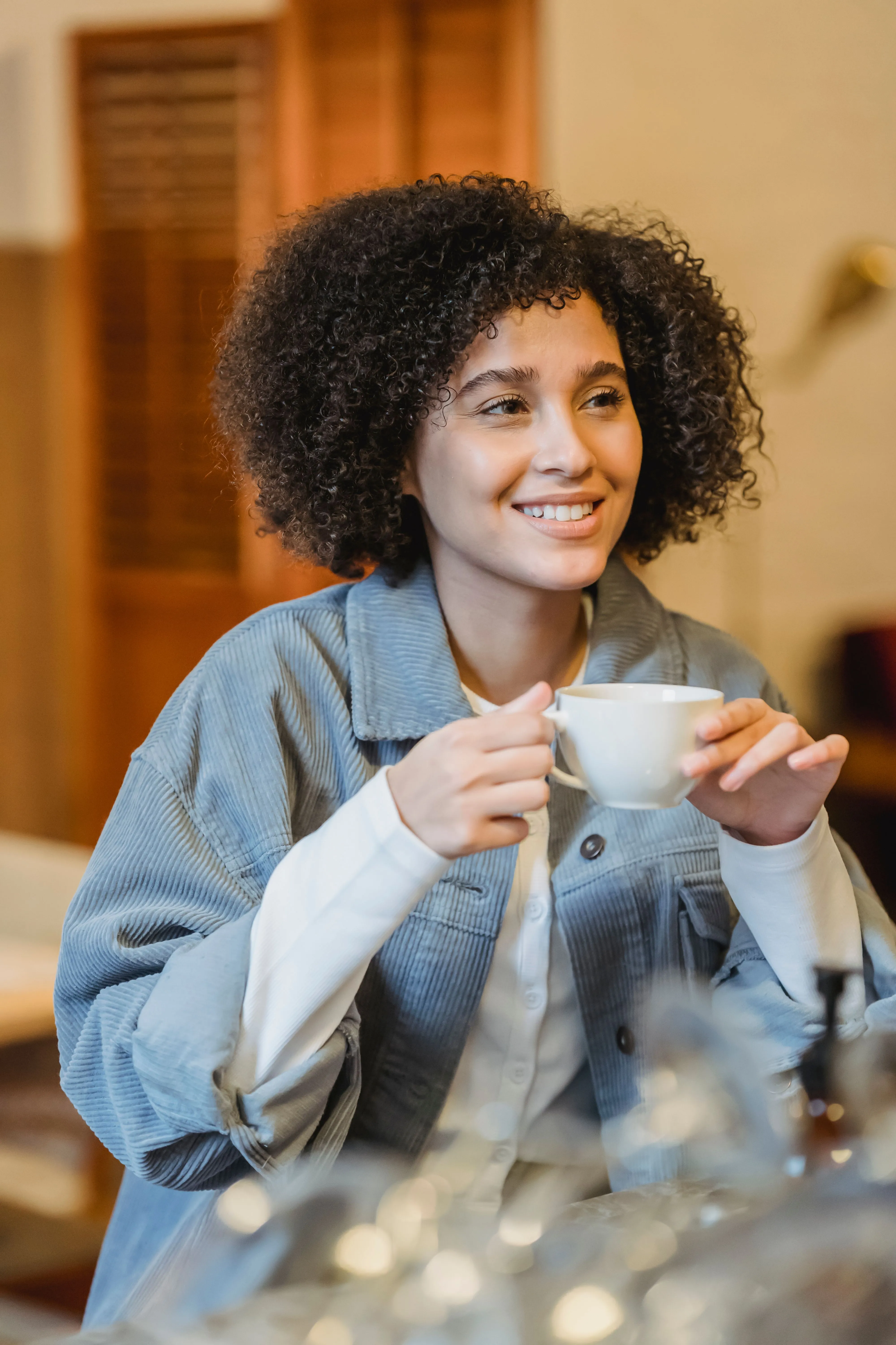 A woman with curly hair holding a cup of coffee, smiling at something across the room