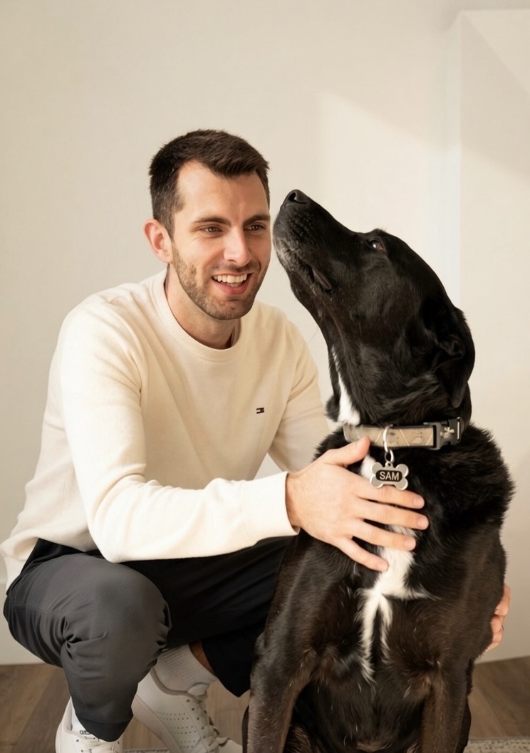 Mark crouching and laughing with Sam, a black dog with a white chest, who is nuzzling his cheek