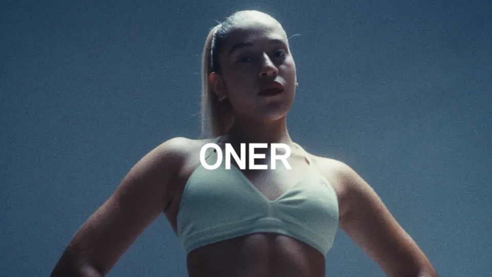 Low-angle shot of Angelina, hands on her hips in a mint Oner sports bra against a blue-grey wall, with “ONER” in white — This Is How I Oner My Body, a brand film by MindWalk Pictures