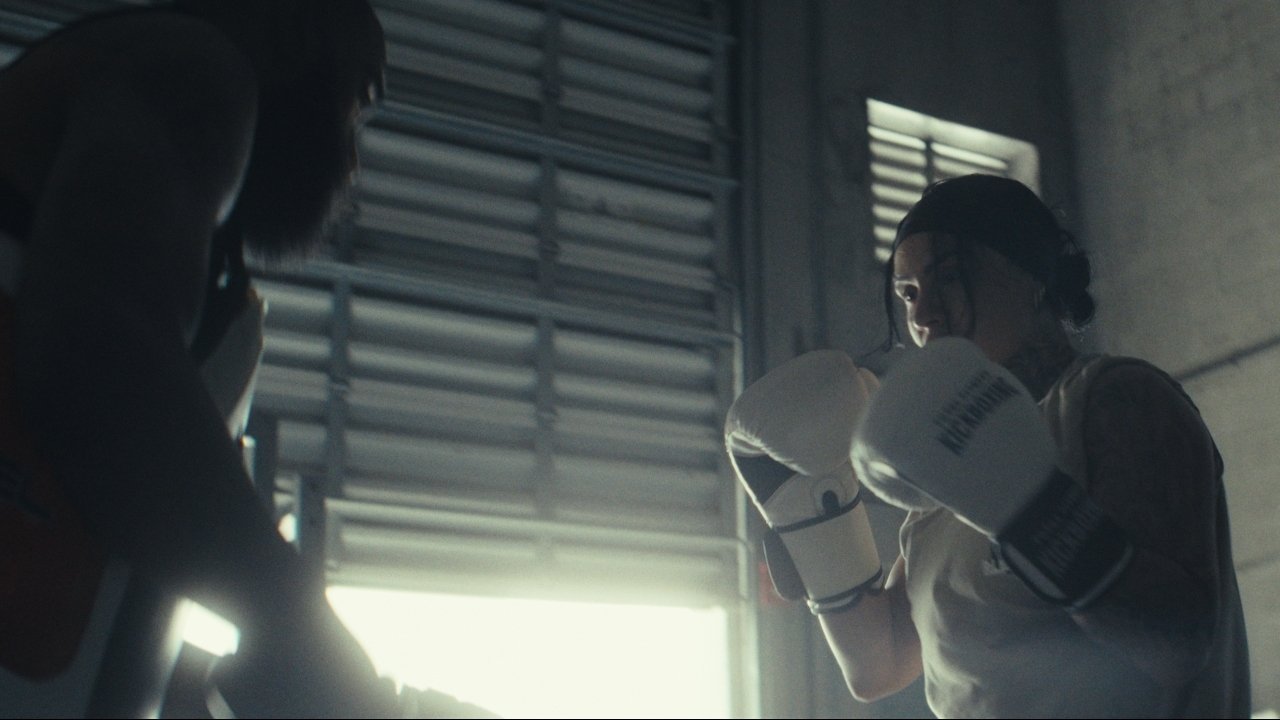 Cassidy with her gloves up, facing her coach in a dim gym lit by a bright shuttered window — Inconquerable, a brand film by MindWalk Pictures