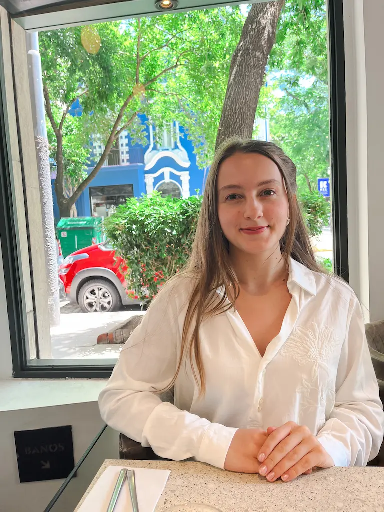 Young woman with long brown hair, wearing a white button-up shirt, sitting at a table with folded hands in front of a large window showing green trees and a blue building outside.