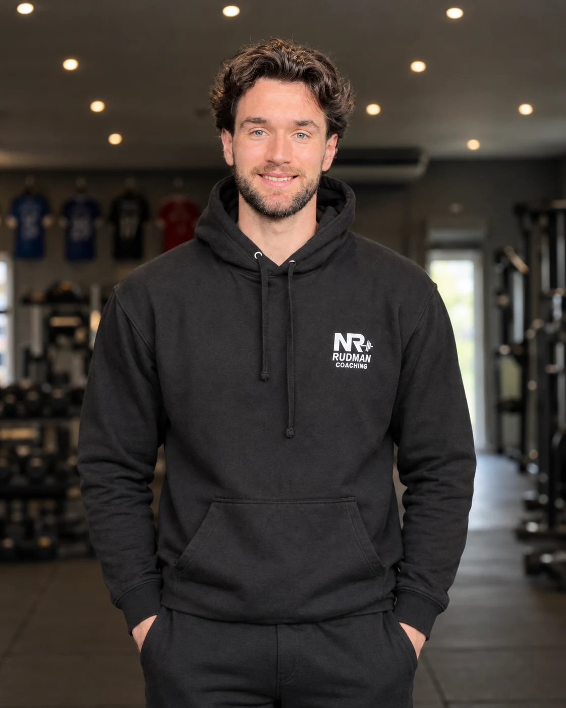 Nathan Rudman, personal trainer, standing in the gym in a black Rudman Coaching hoodie