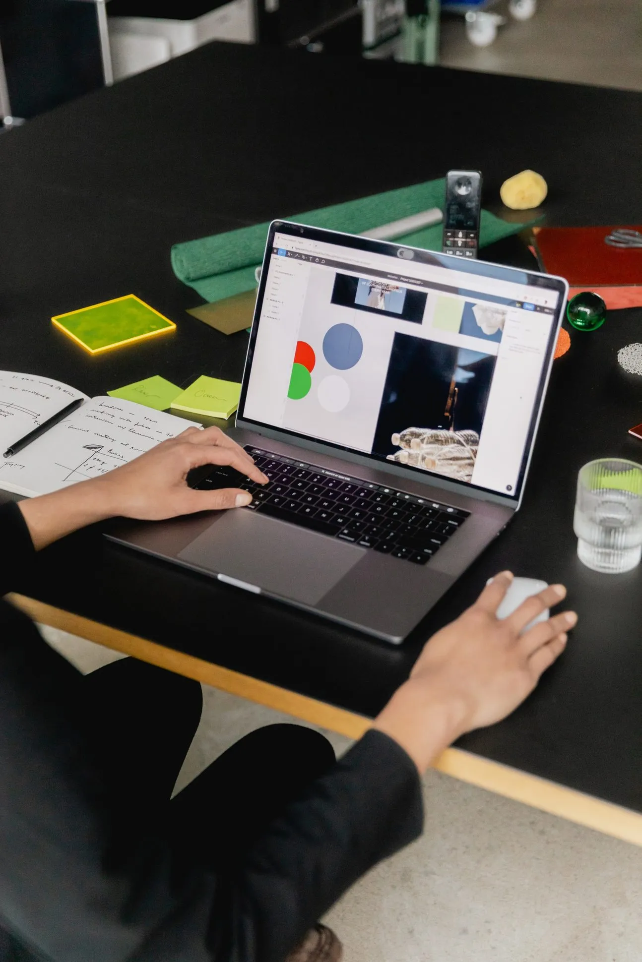 Hands on a laptop showing a design layout, next to a handwritten notebook and sticky notes