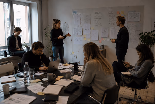 Office meeting with five people in a modern workspace; two are standing near a wall covered with papers and sticky notes discussing ideas while three sit at a cluttered table with laptops, notebooks, and cups, engaged in work.