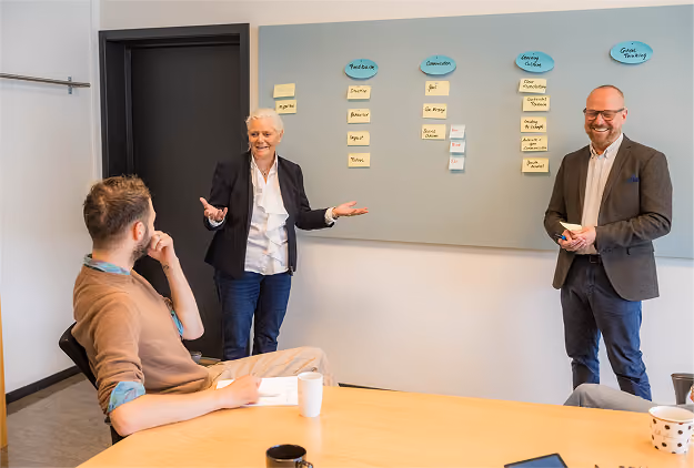 Three business people in a meeting room; two of them stand by a whiteboard with sticky notes under headers labeled Feedback, Discussion, Evening Actions, and Goal Tracking, while the third person sits at a table with a notebook and mug, listening and engaging.