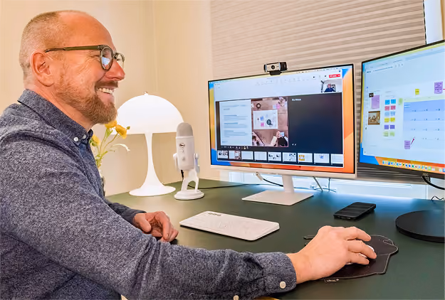 A man with glasses and a beard smiling while sitting at a desk using a mouse in front of two computer monitors, one showing a video call and the other displaying a calendar with sticky notes. A white microphone and a lamp are also on the desk.