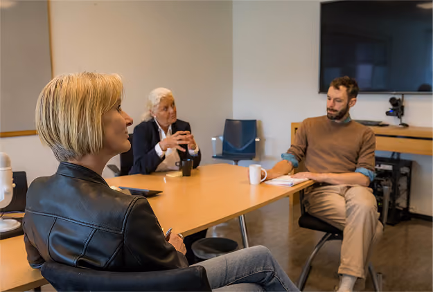 Three people sitting around a conference table engaged in a discussion: a woman with short blonde hair in a black jacket in the foreground, an older woman with white hair and a dark blazer in the middle, and a man with a beard and brown sweater holding a coffee mug on the right.
