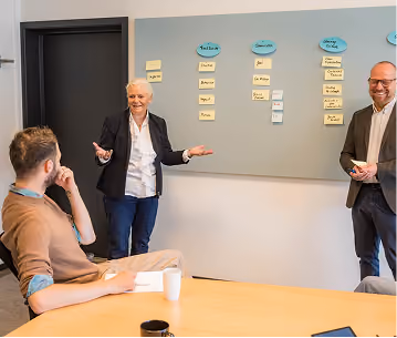 A woman in a black blazer stands near a whiteboard with sticky notes, speaking to two seated men in a meeting room.