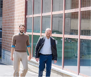 Two men walking and talking outdoors alongside a large glass window, with a brick wall in the background.