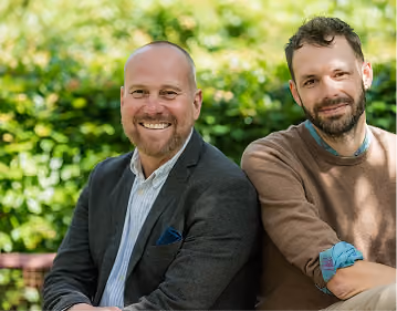 Two men sitting outdoors in front of green foliage; the man on the left is bald with a beard wearing a dark blazer and white shirt smiling, and the man on the right has short curly hair and a beard wearing a brown sweater over a blue shirt smiling slightly.