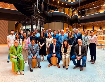 Group photo of 22 diverse people in business attire posing indoors in a modern building with multi-level glass railings and warm lighting, some seated on stools and others standing behind them.