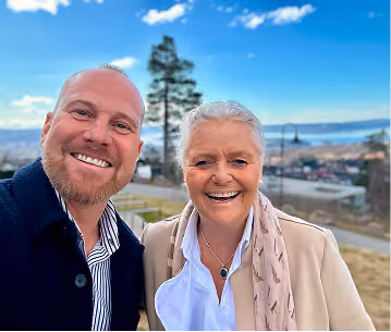 Smiling man and woman outdoors with blurred cityscape and blue sky with clouds in the background.