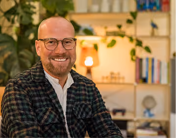 Smiling bald man with glasses wearing a checkered jacket and white shirt sitting indoors with blurred bookshelf and plant in the background.