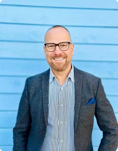 Smiling man with glasses and a beard wearing a dark blazer over a striped shirt, standing in front of a blue wooden wall.