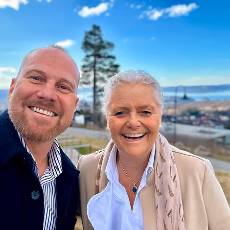 Smiling man and woman outdoors with a clear blue sky, tree, and distant landscape in the background.