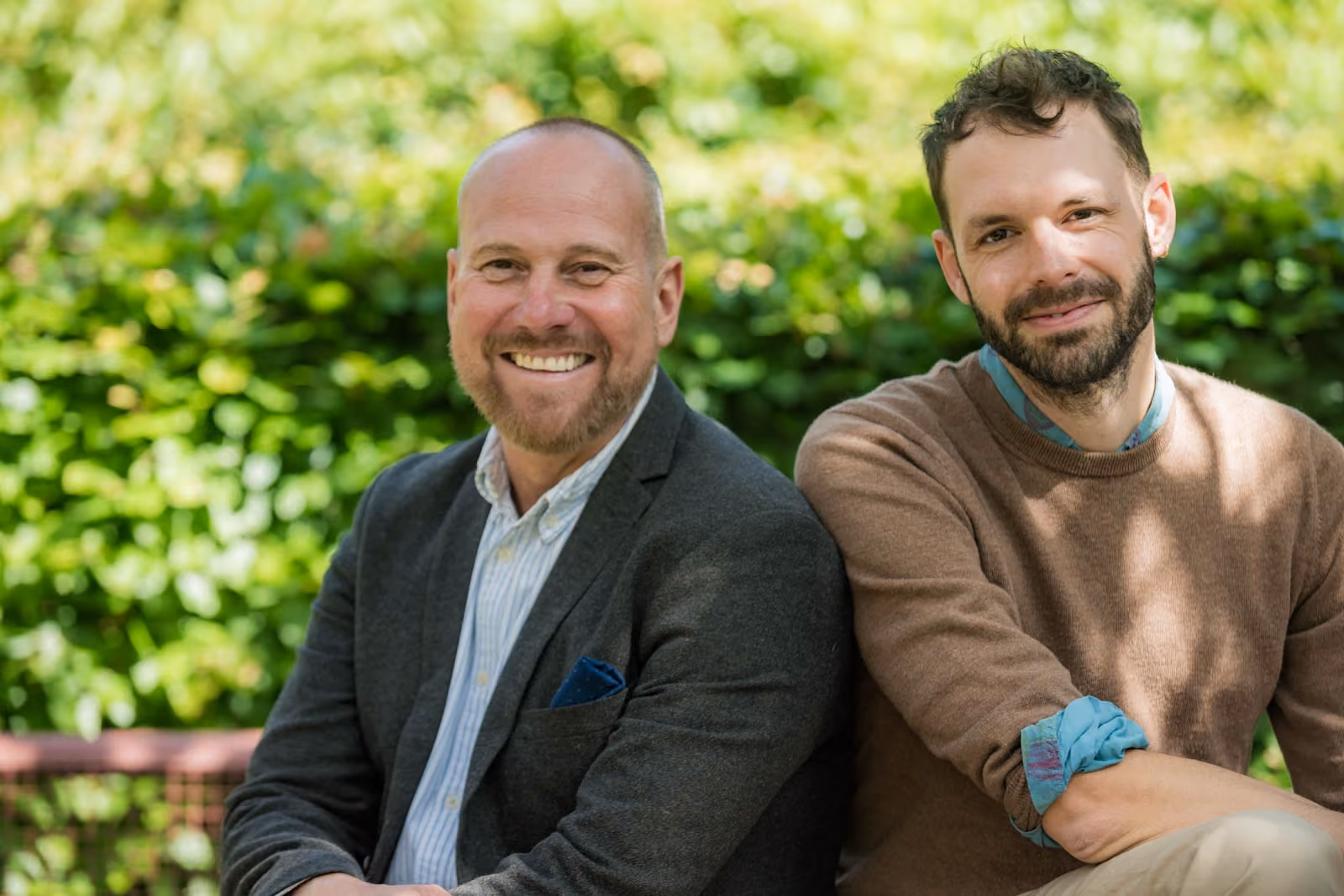 Two men sitting closely on a bench outdoors with green foliage in the background, both smiling at the camera; the man on the left is bald, wearing a dark blazer and light shirt, and the man on the right has short dark hair, a beard, and is wearing a brown sweater with a blue shirt underneath.