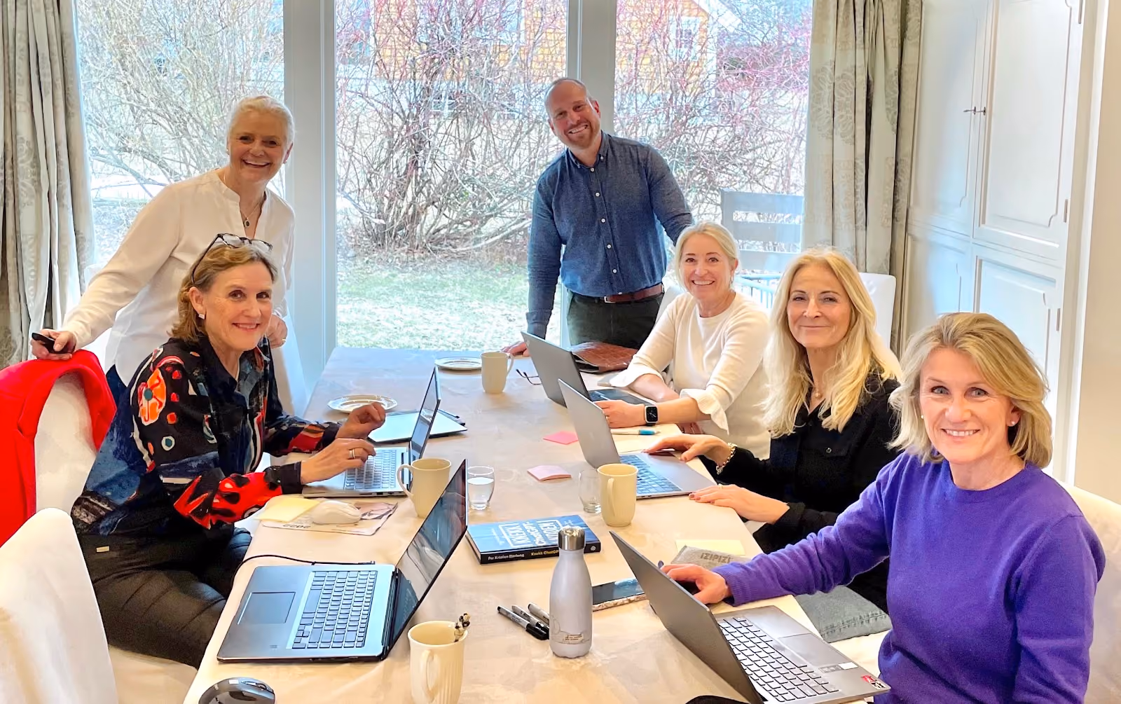 Five people around a table with laptops and mugs, smiling and working together in a bright room with large windows showing a garden outside.