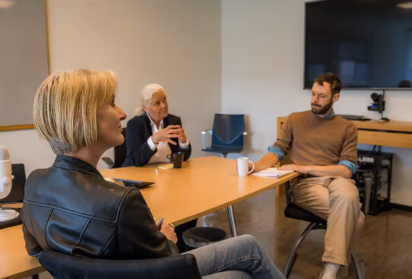 Three people having a discussion around a wooden conference table in an office, with one woman in a black leather jacket in the foreground, an older woman in a dark blazer, and a man in a brown sweater taking notes.