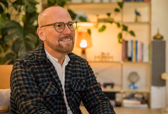 Smiling bald man with glasses and a beard wearing a checkered jacket, sitting indoors with a bookshelf and plants in the background.