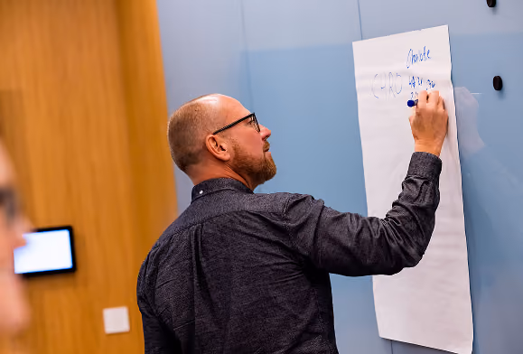 A man with glasses and a beard writing on a large white paper sheet attached to a wall in a meeting room.