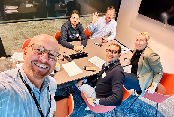 Five people seated around a conference table in a modern office room; one man taking a selfie smiling at the camera, another man waving, and others smiling.