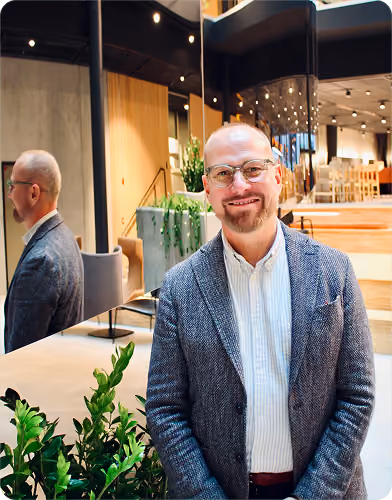 Smiling bald man with glasses and a beard wearing a textured blazer and striped shirt standing indoors next to green plants and a large mirror showing his reflection.