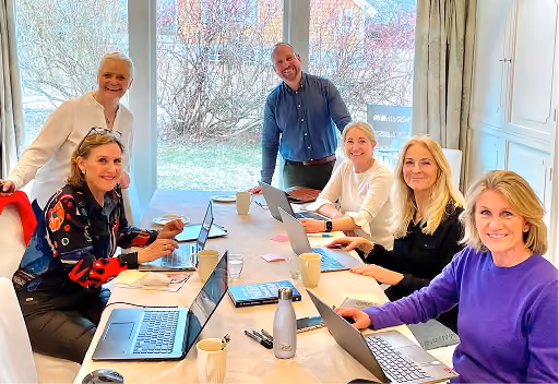 Six colleagues, five women and one man, sitting around a table with laptops, notebooks, and coffee cups in a bright room with large windows, smiling and engaging in a meeting.