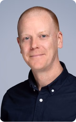 Portrait of a smiling man with short red hair and light skin wearing a dark button-up shirt against a plain gray background.