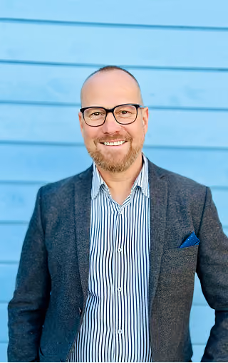 Smiling bald man with glasses, a beard, wearing a striped shirt and a dark blazer with a blue pocket square, standing in front of a blue wooden wall.