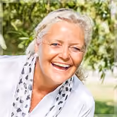 Smiling elderly woman with short gray hair wearing a white top and a scarf with a black and white pattern, outdoors with green foliage in the background.