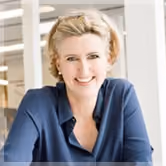 Smiling woman with short blonde hair wearing a dark blue top sitting indoors with blurred background.