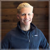 Smiling man with short blond hair wearing a dark zip-up jacket standing in front of a wooden wall.